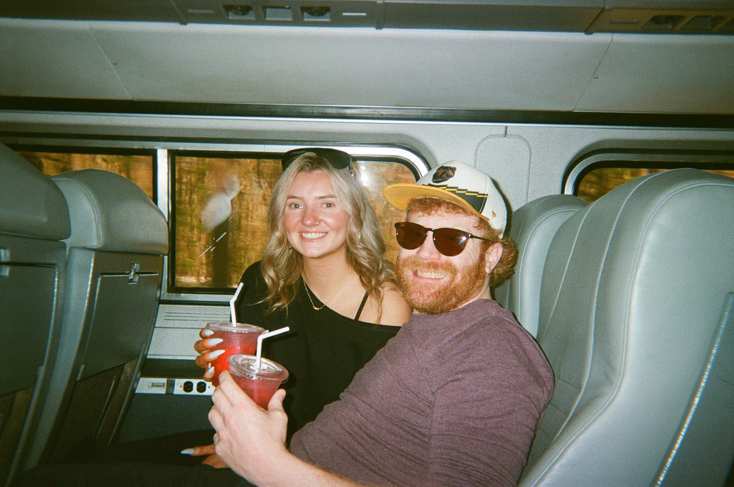 Smiling couple sitting close together on a bus or train, holding red drinks with straws.