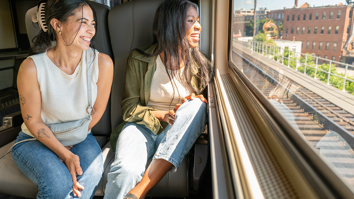 Two friends sit on a train, smiling and looking out the window as sunlight pours in and city buildings pass by.
