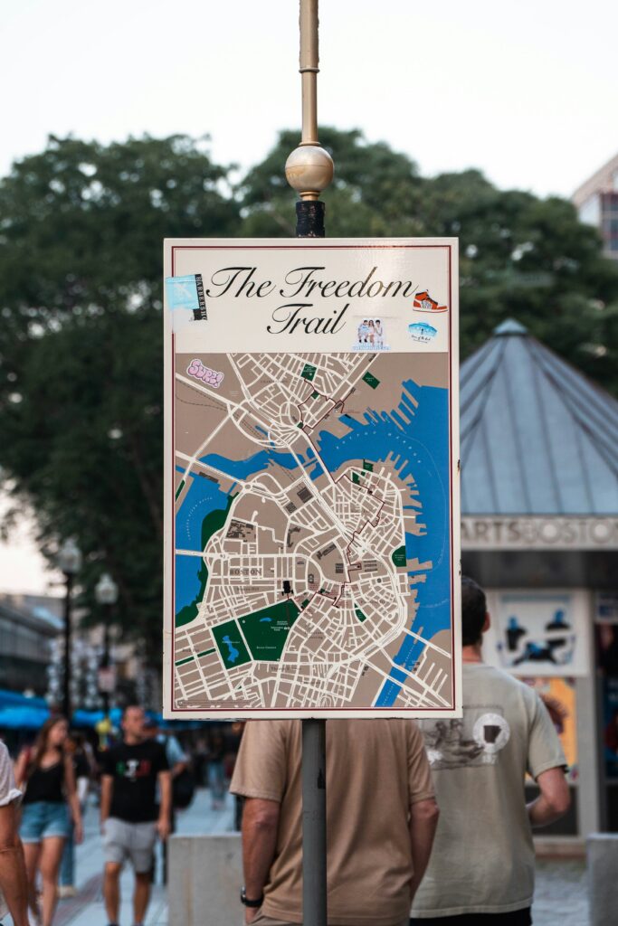 Sign reads The Freedom Trail with a detailed Boston map on a post, stickers around the top, in a busy outdoor setting.